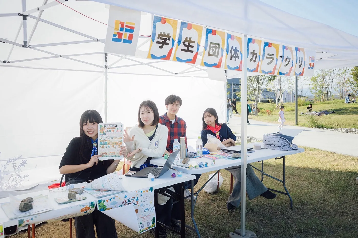 「学生団体カラクリ」の看板を掲げたテントで探検クエストのチラシを見せるメンバー